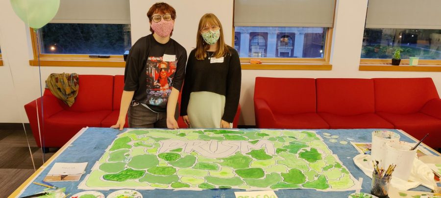 Tosca (left) and Natalie (right) stand behind a sheet of paper covered in painted shades of green. In the middle of the paper in big letters reads Prism.