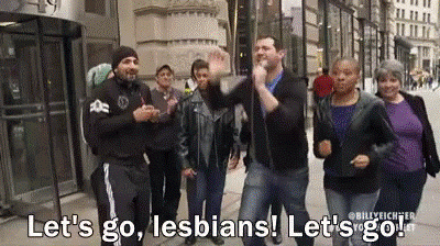 A man is on a sidewalk holding a microphone, followed by a group of people.