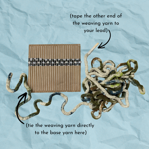 Blue paper texture background with cardboard loom with yarn for weaving, text reads "(tie the weaving yarn directly to the base yarn here) (tape the other end of the weaving yarn to your lead)"