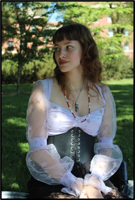 A person sits on the grass in front of some trees and a building.